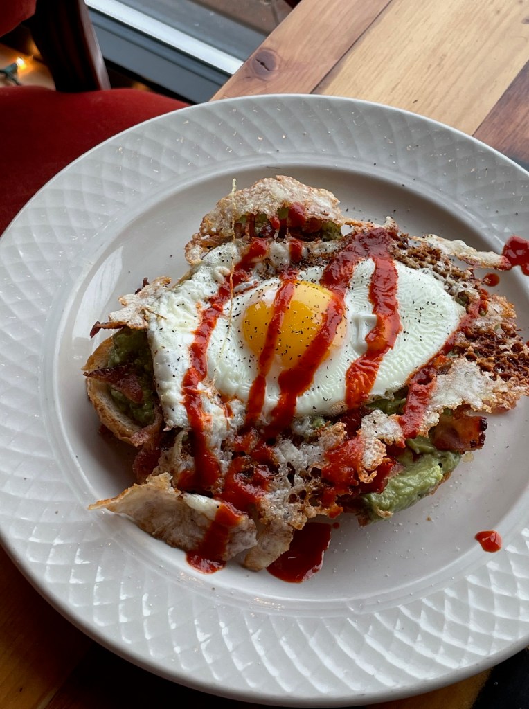 Avocado toast topped with over easy egg, bacon, and salsa roja served during breakfast at neighborhood bar